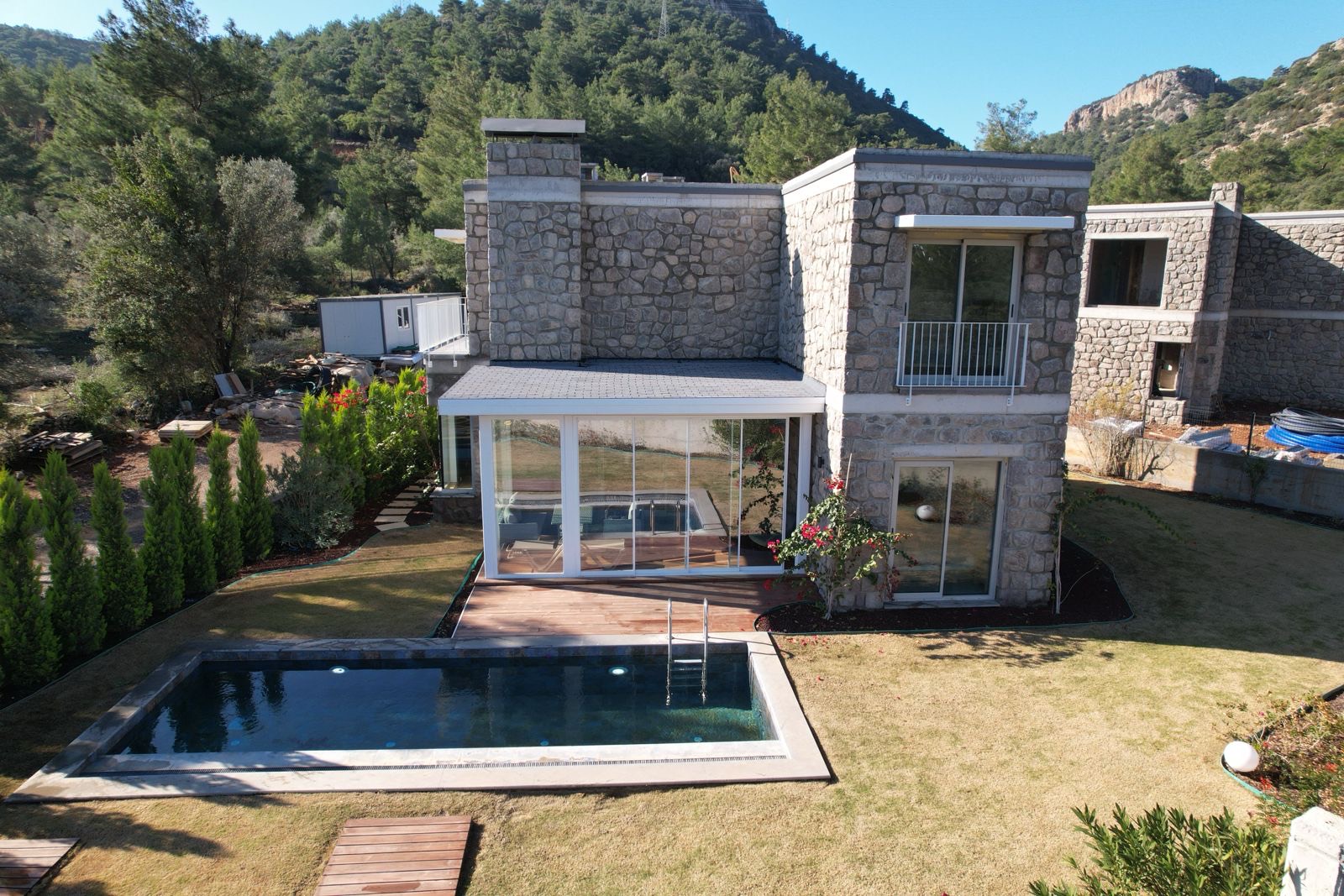 Rinnova Bodrum'da tamamlanmış taş villa; arkasında şantiye konteyneri, malzeme paletleri ve henüz bitmemiş bir komşu blok görünüyor.